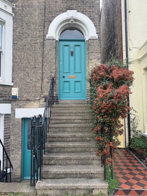 A set of concrete steps leading up to a vibrant turquoise front door with a brass letterbox and door handle, framed by a white arched lintel set into a brown brick building. On the right side of the s